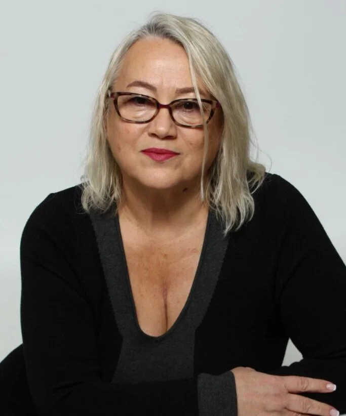 Woman with shoulder-length light blonde hair and tortoiseshell glasses in a black top against a plain grey background